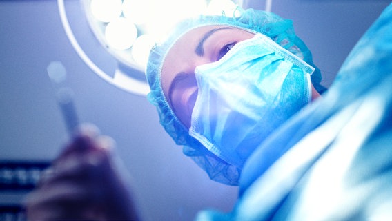 Aus Patientensicht nach oben: mit OP-Maske, Haube, Kittel und Handschuhen bekleidete Ärztin beugt sich mit einem Skalpell in der Hand nach unten. Dahinter eine OP-Lampe. © Addictive Stock / photocase.de 