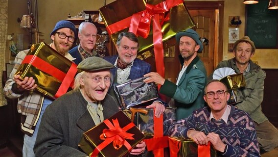 Heinzi, Shorty, Onkel Krischan, Günther Griem, Kuno, Jürgen Seute und Adsche sitzen im Dorfkrug mit Geschenken. © NDR 