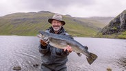 Ein Mann mit Hut steht an einem See und hält lächelnd einen Fisch in seinen Händen. Im Hintergrund eine grüne hügelige Landschaft und tiefhängende Wolken. © NDR/Mario Jeschke Foto: Mario Jeschke