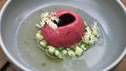 Eine Kugel Himbeereis mit Gurkenstückchen sowie Holunderblüten und -sirup auf einem grauen Teller angerichtet. © NDR 