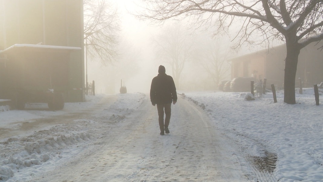 Eine Person läuft im winterlichen Nebel über einen Bauernhof.