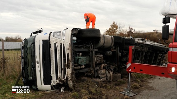 Ein Mann im gelben Arbeitsanzug steht auf einem auf der Seite liegenden Lkw an der A7 zwischen Bordesholm und Warder. © NDR 