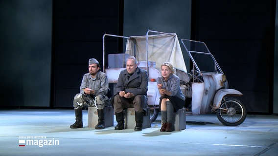 Auf einer Bühne sitzen drei Personen vor einem Kleintransporter, sie zeigen eine Szene aus "Mutter Courage und ihre Kinder". © NDR Foto: NDR Screenhots