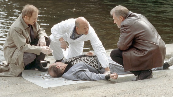 Haustein (Harald Weiler, Mitte) zeigt Eduard Holicek (Tilo Prückner, links) und Jan Casstorff (Robert Atzorn, rechts) seine Beobachtungen an der Leiche von Heinrich Kehl (Jean-Pierre Le Roy). © NDR/Thorsten Jander - Abdruck 