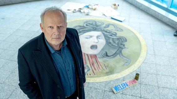 Ein Mann mit grauem Bart und schwarzem Sakko schaut ernst nach oben in die Kamera, hinter ihm ist ein Haupt einer Medusa auf den Boden gemalt - Szene aus dem Tatort "Borowski und das Haupt der Medusa". © NDR/Thorsten Jander Foto: NDR/