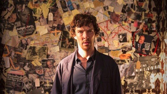 Sherlock (Benedict Cumberbatch) schaut verwirrt ins Leere. Sein Gesicht ist verschwitzt, hinter ihm sind die Wände mit zahlreichen Zeitungsartikeln beklebt. © ARD Degeto/BBC / Hartswood Films 2016 