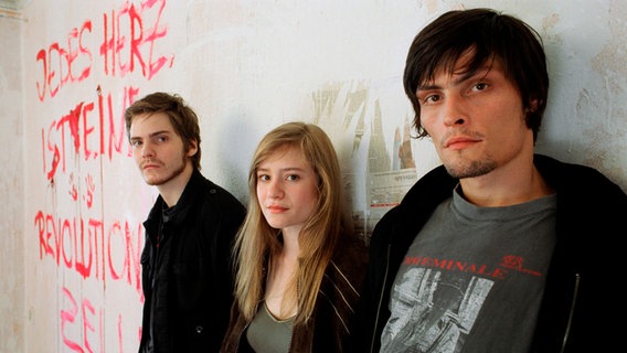 V.li.n.re.: Jan (Daniel Brühl), Jule (Julia Jentsch) und Peter (Stipe Erceg) stehen an einer Wand gelehnt von einer Zimmerwand. Die Wand scheint renovierungsbedürftig, mit roter Farbe steht darauf geschrieben "Jedes Herz ist eine revolutionäre Zelle". © Bild: SWR/y3 film 