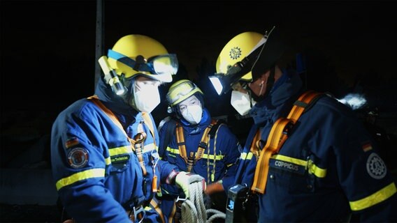 Drei Mitarbeitende des THW mit Schutzkleidung und -masken holen beim Einsatz ein Seil. © NDR/Kinescope Film 
