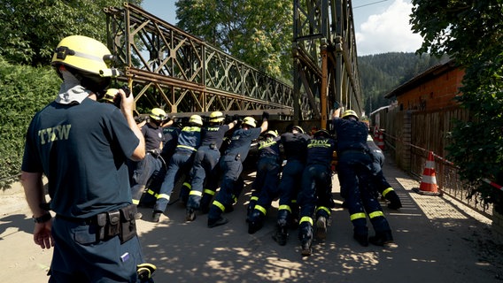 Mitarbeitende des THW drücken gemeinsam gegen den Teil einer Brücke. © NDR/Kinescope Film 