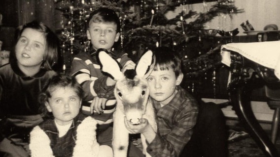 Eine historische schwarz-weiß Fotografie zeigt Kinder mit einem Stofftier vor einem Weihnachtsbaum.  