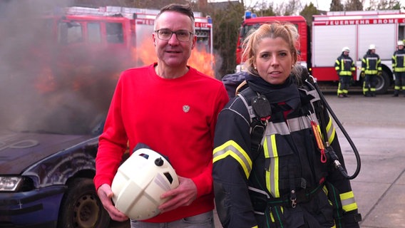 Reporter Torben Hildebrandt und Feuerwehrfrau Theresa Balzer stehen vor einem brennenden Fahrzeug. © NDR 