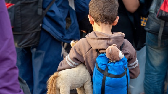 Ein Junge mit Rucksack und Stofftieren von ist von Hinten in einer Gruppe von Erwachsenen zu sehen. © fotolia.com Foto: Lydia Geissler