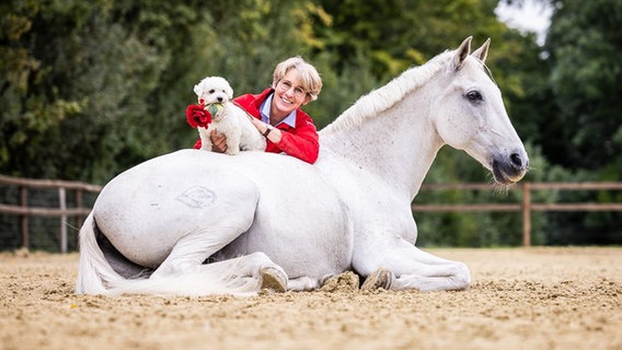 Tier-Trainerin Anne Krüger-Degener ist an ein weißen Schimmel angelehnt, mit einem kleinen weißen Hund auf dem Pferderücken © Anna Auerbach Foto: Anna Auerbach