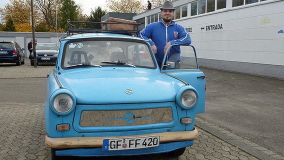 Ein Mann steht in DDR-Trainingsjacke und Hut an einem hellbaluen Trabbi. © NDR Foto: Peter Jagla