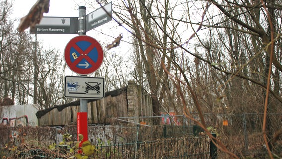 Hinter den Schildern "Berliner Mauerweg", die auf einen Knick im Verlauf der ehemaligen Grenze hinweisen, ist eine Mauer mit Stacheldraht zu erkennen. © NDR Foto: Daniel Sprenger