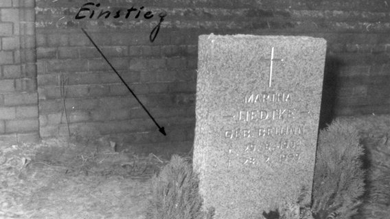 Historische Aufnahme vom Friedhof Pankow, hinter einem Grabstein vor der Friedhofsmauer ist ein auf das Bild aufgemalter Pfeil zu sehen, der auf ein Loch deutet, darüber steht "Einstieg". © BStU 