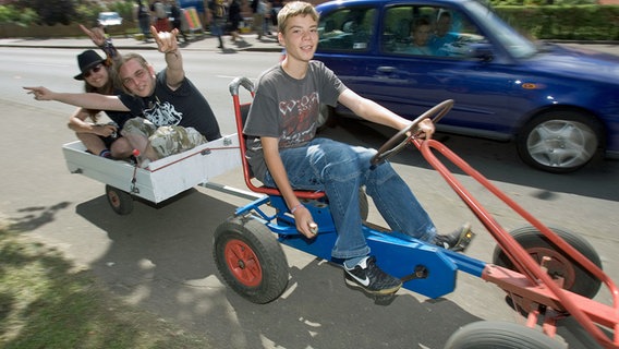 Zwei Heavy-Metal-Fans lassen sich am 3. August 2007 mit einem Kettcar über einen Bürgersteig in Wacken bis zum Festivalgelände ziehen. © picture alliance/dpa Foto: Friso Gentsch