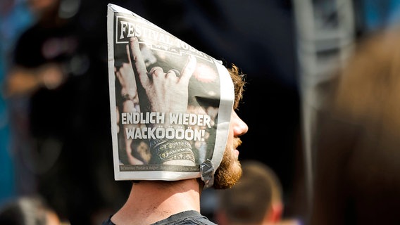 Ein Metal-Fan trägt eine Zeitung als Sonnenschutz mit dem Titel "Endlich wieder Wacköööön!" (3. August 2022). © picture alliance/dpa Foto: Frank Molter