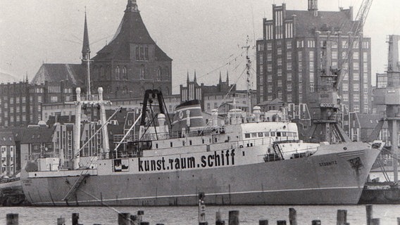 Die "Stubnitz" liegt 1993 im Hafen von Rostock, hinter sind Häuser in Backsteinoptik zu sehen: Auf der Flanke des Schiffs prangt die Aufschrift "kunst.raum.schiff" © Stubnitz Archiv 