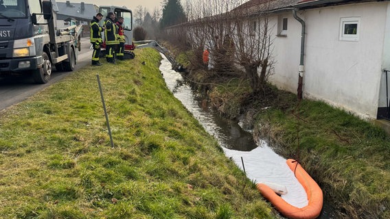 Auf einem Bach schwimmt eine orange Ölsperre. Davor hat sich weißes Ölbindemittel gesammelt. im Hintergrund stehen Feuerwhrleute an einem Feuerwehrfahrzeug. © Feuerwehr Gemeinde Kalefeld Foto: Tim Brand