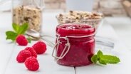 Ein Glas mit Himbeermarmeldae neben frischen Himbeeren auf einem weißen Holztisch mit Müsligläsern im Hintergrund.. © fotolia.com Foto: Fischer Food Design