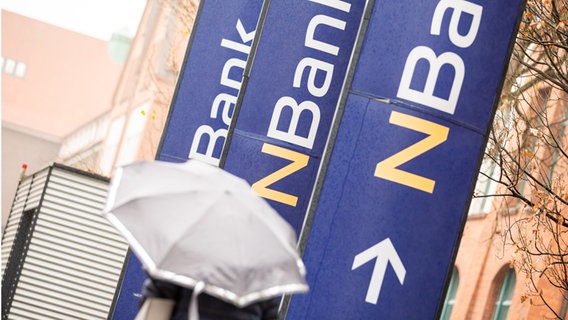 Eine Frau mit Regenschirm geht an Werbepylonen mit dem "NBank"-Logo am Sitz der "NBank", der Investitions- und Förderbank des Landes Niedersachsen, vorbei. © dpa-Bildfunk Foto: Moritz Frankenberg/dpa