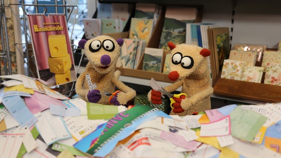 Zwei Erdmännchen aus Stoff sitzen hinter einem Papierberg aus zerschnittenen Kalendern. Beide haben eine Schere in der Hand. © bigSmile/NDR 