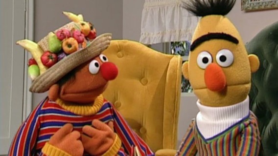 Ernie und Bert sitzen in ihren Sesseln. Ernie trägt einen Hut mir Früchten © NDR 