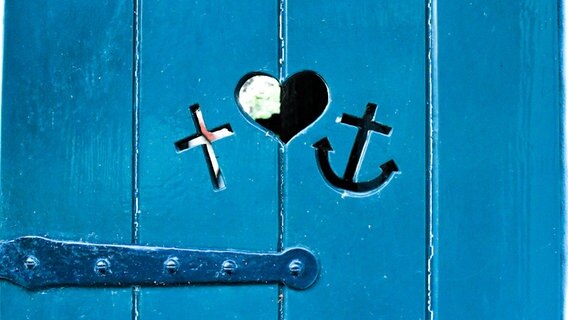 Ein Kreuz, ein Herz und ein Anker in eine Holztür geschnitzt. © picture alliance / Zoonar Foto: Eva-Maria Pollich