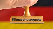 Ein Holzstempel mit der Aufschrift "Klimaschutzgesetz", gehalten von einer Hand im Anschnitt vor einer unscharfen Deutschland-Flagge im Hintergrund. © picture alliance / SULUPRESS.DE Foto: Torsten Sukrow/SULUPRESS.DE