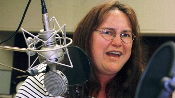 Eine langhaarige Frau mit Brille steht in einem Studio vor einem Mikrofon und spricht hinein. Sie trägt ein weiß blau gestreiftes Sweat Shirt und blickt in die Kamera. © NDR 