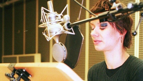 Eine kurzhaarige Frau steht in einem Studio vor einem Mikrofon und spricht hinein während sie von einem Pult den Text abliest. © NDR 