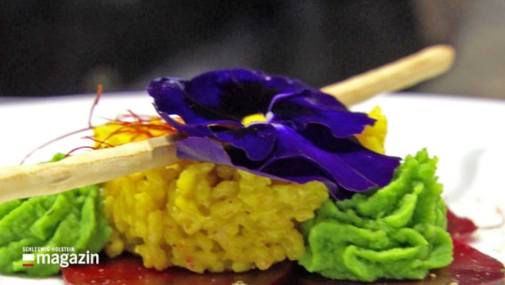 Ein auf dem Teller auf Rote Bete-Carpaccio Safran-Risotto, das mit einer blauen Blume dekoriert ist. © NDR 