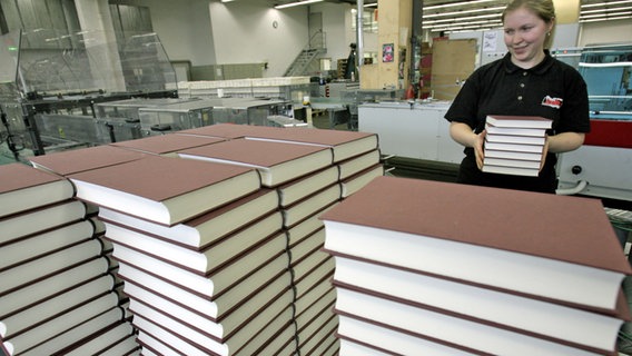 Eine Mitarbeiterin hält gedruckte Bücher in der Hand, vor ihr und um sie herum lagern hohe Bücherstapel - ohne Aufschrift und ohne Cover © picture-alliance/ dpa | Jan-Peter Kasper Foto: Jan-Peter Kasper