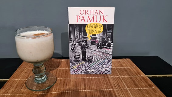 Ein Buch von Orhan Pamuk steht neben einem Glas türkischem Boza mit Zimt bestreut - Bild zum Podcast eat READ sleep  Folge 28 Türkisches Boza mit David Safier © NDR Foto: Jan Ehler
