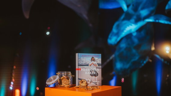 Das Buch "The Hebridean Baker" von Coinneach MacLeod steht auf einem orangefarbenen Tisch, daneben liegen Kekse © NDR / marcusfriedrich.media Foto: Marcus Friedrich