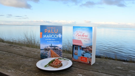Ein Krabbenbrot liegt neben zwei Büchern auf einem Teller. Im Hintergrund sieht man das Meer. © NDR/Manfred Koch Foto: Manfred Koch
