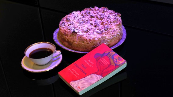 Eine Tasse Kaffee, ein Kuchen und ein Buch sind nebeneinander arrangiert. © NDR 