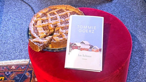 Ein weißes Hardcoverbuch mit dem Titel "Im Schnee" lehnt an einem runden Kuchen, beides steht auf einem roten Samthocker © NDR Foto: Katharina Mahrenholtz