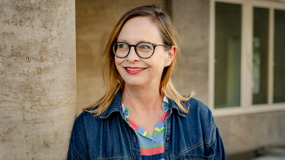 Eine Frau mit Brille und Jeansjacke steht an eine Wand gelehnt und lächelt. © Patricia Haas 