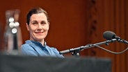 Eine Frau (Nina Kunzendorf) sitzt auf einem Podium mit Mikrofon und lächelt. © IMAGO / Future Image 