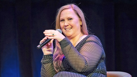 Eine blonde Frau mit langen Haaren und dunkelblauem Kleid mit weißen Pünktchen hält lächelnd ein Mikro in der Hand, an dem einen Finger trägt sie einen goldenen Ring mit grünem Stein - Bestsellerautorin Rebecca Yarros bei einer Lesung in New York © picture alliance / CJ Rivera/Invision/AP | CJ Rivera Foto:  CJ Rivera