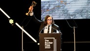 Ein Mann mit schwarzem Anzug, weißem Hemd, schwarzer Krawatte hält einen goldenen Preis in der Hand hoch - Paul Lynch gewinnt den Booker-Preis © Alberto Pezzali/AP/dpa +++ dpa-Bildfunk +++ Foto: Alberto Pezzali