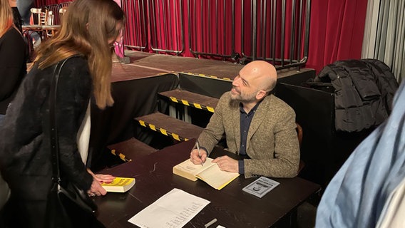 Roberto Saviano sitzt im braunen Sacko an einem Tisch und signiert Bücher, dabei schaut er lächelnd zu einer Frau hoch, die vor dem Tisch steht. © Jens Büchsenmann / NDR Foto: Jens Büchsenmann / NDR