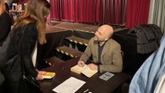 Roberto Saviano sitzt im braunen Sacko an einem Tisch und signiert Bücher, dabei schaut er lächelnd zu einer Frau hoch, die vor dem Tisch steht. © Jens Büchsenmann / NDR Foto: Jens Büchsenmann / NDR