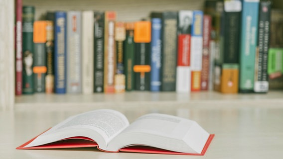 Ein aufgeklapptes Buch liegt auf einem Tisch, im Hintergrund schemenhaft weitere Bücher in einem Regal. © picture alliance / Zoonar Foto: Oleksandr Latkun