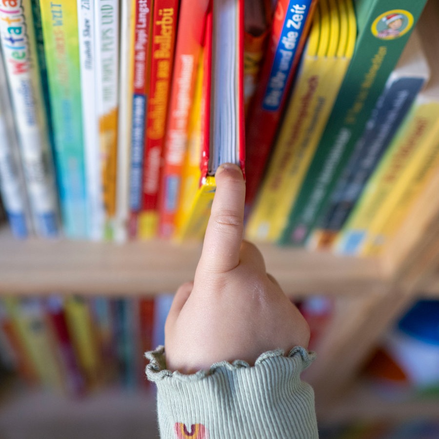 Eine Kinderhand zieht ein Buch aus einem Bücherregal. © picture alliance/dpa | Hendrik Schmidt 