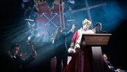 Ein als antiker Herrscher inszenierter Mann auf einer dunklen Bühne mit erleuchtetem Kreuz im Hintergrund in Jelineks Stück "Endsieg" im Hamburger Schauspielhaus. © Thomas Aurin 