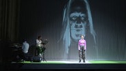 Eine Frau steht auf einer Bühne, im Hintergrund sitzen zwei Musiker © Screenshot NDR 