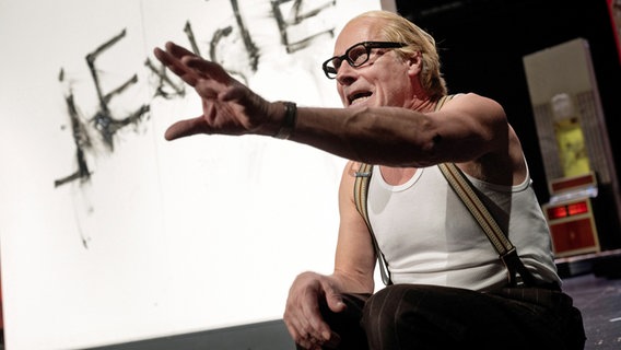 Ein älterer Mann (der Schauspieler Wolfram Koch) mit Brille, in Unterhemd mit Hosenträgern, sitzt auf der Bühne und hat den linken Arm erhoben. © Armin Smailovic/Schauspielhaus Zrich Foto: Armin Smailovic